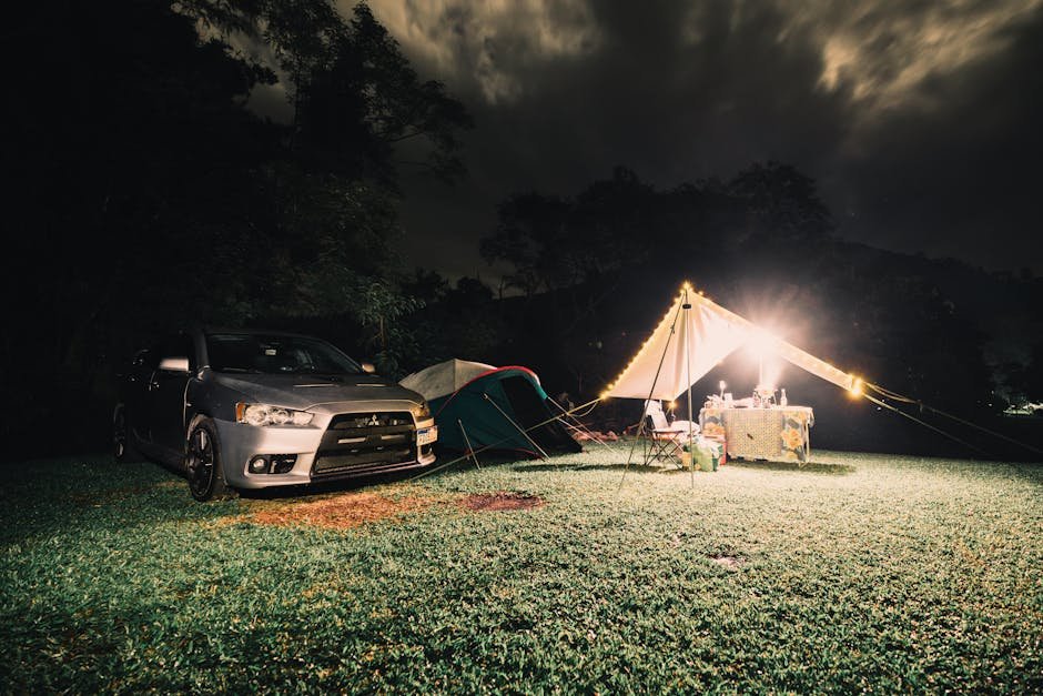 Serene camping site at night featuring illuminated tents, car, and lush grass under a cloudy sky.